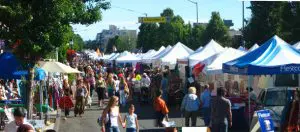A crowded outdoor street market with rows of white tents, vendors, and shoppers on a sunny day.