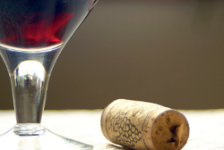 A close-up of a glass of red wine next to a wine cork lying on a light surface.