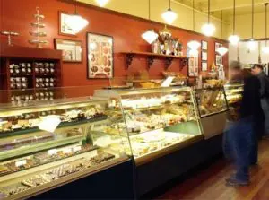 A bakery display case filled with assorted pastries and desserts, with warm lighting and a few customers visible in the shop.
