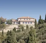 A large, two-story house with a tiled roof sits on a hill surrounded by trees and shrubs under a clear blue sky.