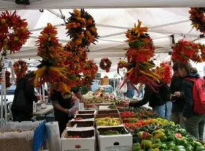 Outdoor market stall with colorful hanging chili pepper arrangements, boxes of assorted vegetables, and people browsing under a white canopy.