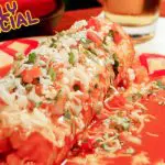 A cheesy burrito topped with salsa and cilantro on a colorful plate, with a “Daily Special” sign in the corner and a drink in the background.