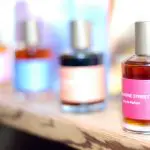 Four glass perfume bottles with colorful labels and silver caps are lined up on a wooden surface, with the focus on the bottle in the foreground labeled "MAGAZINE STREET.