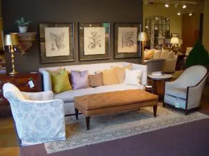 A living room showroom features a beige sofa with multiple cushions, two armchairs, a tan ottoman, a rug, wall art, and various decorative items.