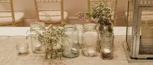 Several glass mason jars with small flowers and tealight candles arranged on a burlap runner, beside a metal lantern, on a decorated table.