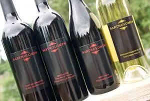 Four bottles of Bartholomew wine, three red and one white, are lined up outdoors. The labels display different varietals and years from Columbia Valley and Walla Walla Valley.