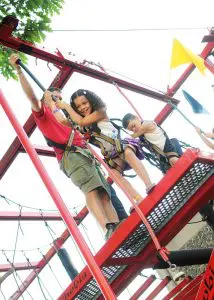 Three people wearing harnesses stand on a red outdoor climbing structure; an adult assists two children as they prepare to climb.