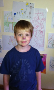 A young boy with marker scribbles on his face stands in front of a wall covered with children's drawings.