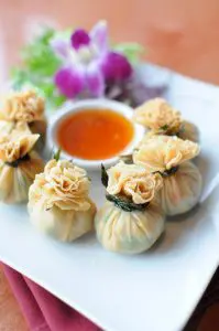 Five golden fried dumplings tied with seaweed, arranged in a circle on a white plate with a small bowl of dipping sauce and a purple orchid garnish.