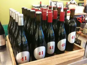 bin 41 anniv A wooden crate filled with wine bottles labeled "41," featuring both red and silver caps, sits on a countertop with office supplies in the background.
