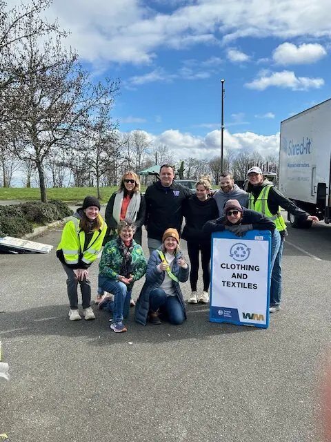 A group of nine people pose outdoors near a "Clothing and Textiles" donation sign, wearing casual and safety vests, with a truck and trees in the background.