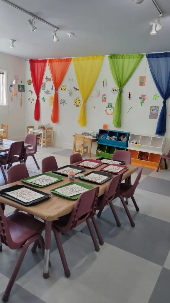 A classroom with small tables and chairs, art trays with tree drawings, colorful curtains, wall art, and shelves with children's supplies.