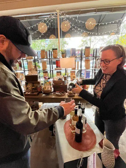 A woman pours wine into a glass for a man at an indoor tasting event, with bottles and gift items displayed on a table and shelves behind them.