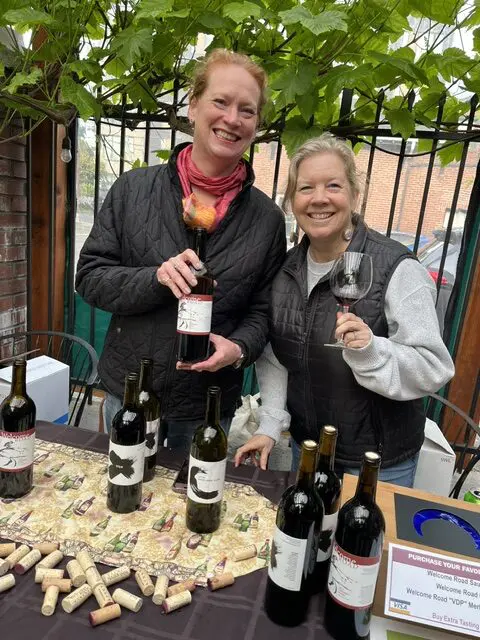 Two women stand behind a table with several wine bottles and corks, one holding a wine bottle and the other holding a wine glass, at an outdoor wine-tasting event.