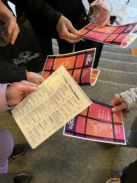 Four people stand on carpeted stairs holding wine walk tasting menus and glasses, comparing selections.