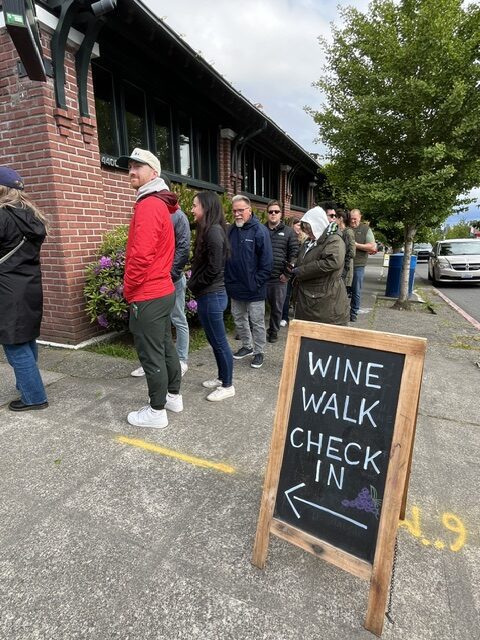 People stand in line outside a brick building next to a sidewalk sign that reads "Wine Walk Check In" with an arrow pointing left.