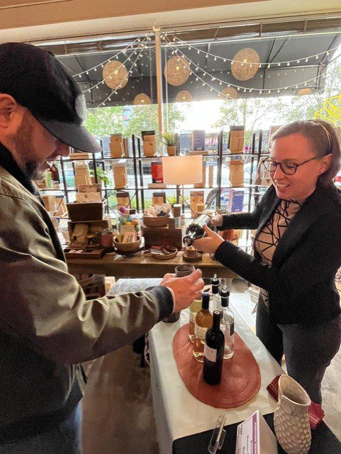 A woman pours wine into a glass for a man at an indoor tasting event, with bottles and gift items displayed on a table and shelves behind them.