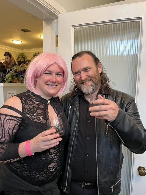Two people stand indoors, smiling at the camera and holding drinks. One wears a pink wig and black lace dress; the other wears a black shirt and leather jacket.