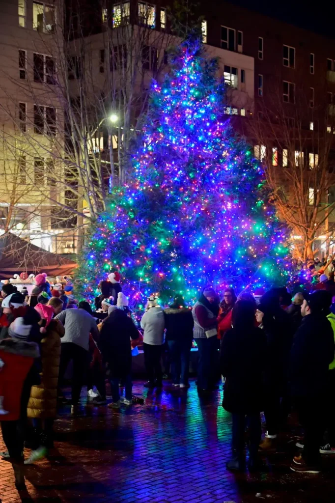 A large crowd gathers around a brightly lit Christmas tree with multicolored lights in an outdoor urban setting at night.
