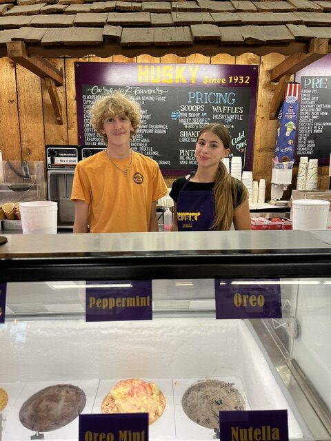 Two employees stand behind the counter at an ice cream shop, with tubs of ice cream and a menu board visible in the background.