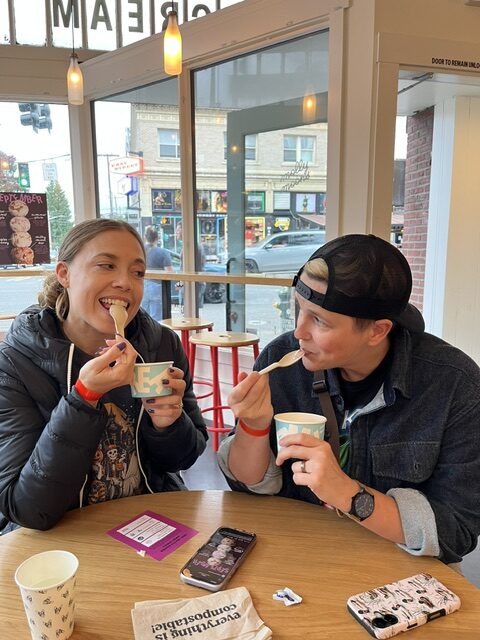Two people sit at a round table in an ice cream shop, smiling and eating ice cream with spoons. Their phones and napkins are on the table. Large windows show a street view outside.