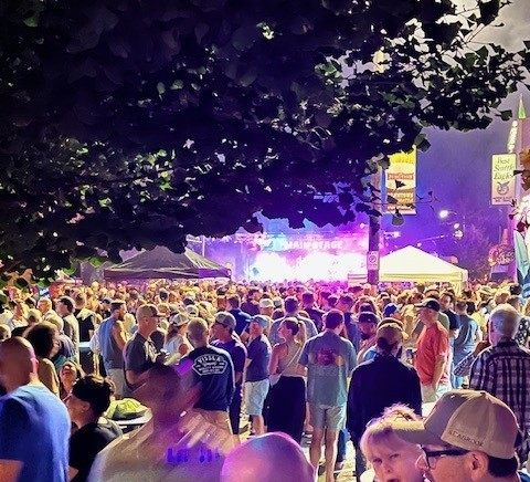 Large crowd gathered outdoors at night in front of a brightly lit stage, with trees and vendor tents visible in the background.