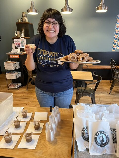 A person smiles while holding a tray of donuts and a sample cup in a café, with stacks of pastries and pudding cups arranged on the table in front of them.