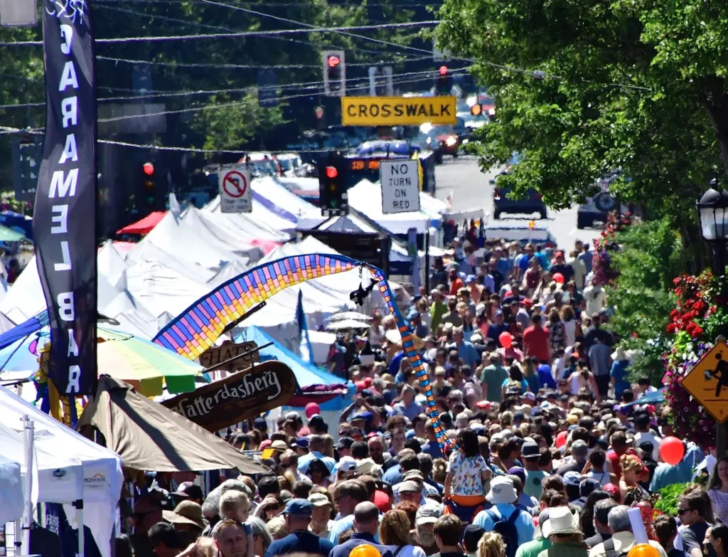 A large crowd walks through an outdoor street festival lined with vendor tents. Colorful decorations and signs are visible above the people. Trees and street signs appear in the background.