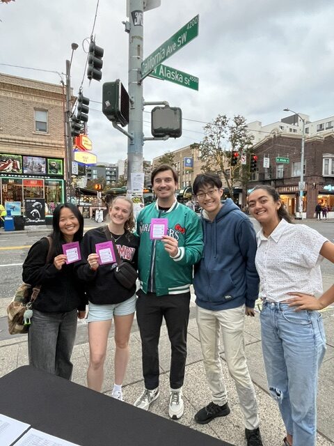 Five people stand on a city street corner holding pink cards, smiling at the camera. Street signs for California Ave SW and Alaska St are visible above them.