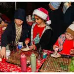 Three children decorate cookies at a table covered with red and white cloth, using frosting bottles. The middle child wears a Santa hat; the others wear winter hats and jackets.
