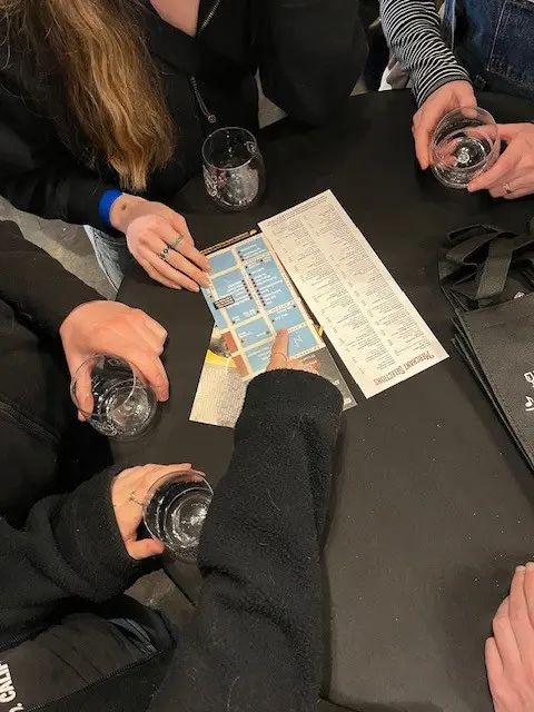 Five people sit around a table, each holding a clear glass, and look at a printed menu or map placed at the center.