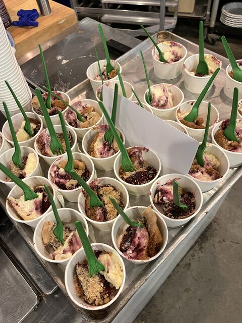 A metal tray holds many paper cups filled with dessert, each with a green plastic spoon and topped with crumbles and berry sauce.