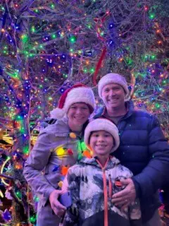 Three people wearing Santa hats stand smiling in front of colorful Christmas lights and decorated trees.