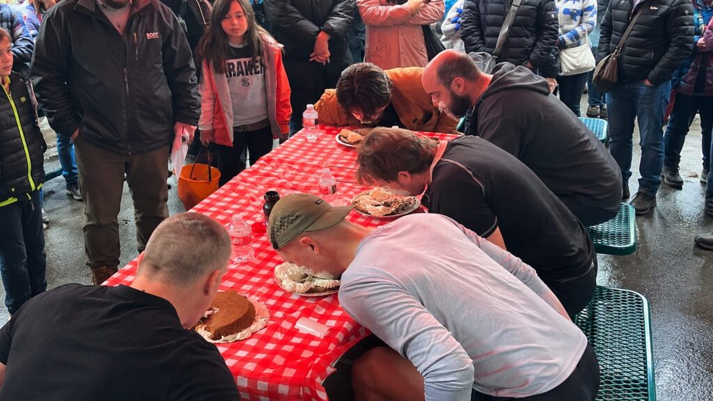 Several people participate in a pie-eating contest at a red checkered table, surrounded by a crowd of onlookers in outdoor clothing.