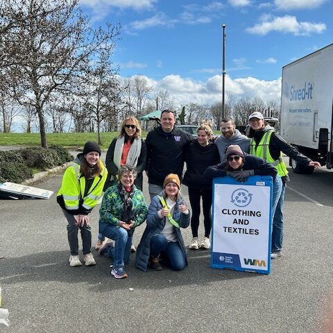 A group of nine people pose outdoors near a "Clothing and Textiles" donation sign, wearing casual and safety vests, with a truck and trees in the background.